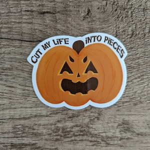 May include: A Halloween-themed sticker featuring an orange pumpkin with a carved face. The sticker has the text "CUT MY LIFE INTO PIECES" in a curved font around the pumpkin. The sticker is on a wooden surface.