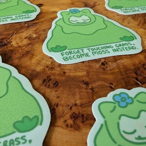 Peut inclure: Plusieurs autocollants repr&eacute;sentant un personnage de dessin anim&eacute; dans un costume de mousse vert avec une fleur bleue. Le texte dit : "Oubliez de toucher l'herbe, devenez de la mousse &agrave; la place."