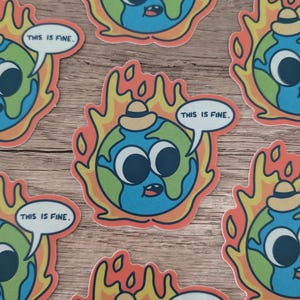 K&ouml;nnte beinhalten: Ein Set mit f&uuml;nf Aufklebern, die eine Karikatur der Erde mit Glotzaugen zeigen, die einen Hut tr&auml;gt und von Flammen verschlungen wird. Jeder Aufkleber tr&auml;gt den Text "This is fine." in einer Sprechblase.