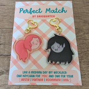 May include: Two acrylic keychains with gold-colored hardware, one featuring a pink-haired character and the other a black-haired character. The keychains are on a pink and white checkered card with the text "Perfect Match" and "Like a modern day BFF necklace".