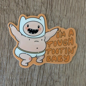 May include: A cartoon sticker of a baby Finn from Adventure Time wearing a diaper and a white hat. The sticker has an orange background with the text "I'm a tough tootin' baby".