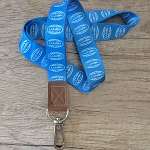 May include: A blue lanyard with the word "LUMON" printed repeatedly in white. The lanyard has a brown leather piece and a silver metal clasp. The lanyard is laying on a wooden surface.