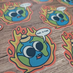 K&ouml;nnte beinhalten: Ein Aufkleber mit einer Cartoon-Erde, die einen Hut tr&auml;gt und in Flammen steht, mit dem Text "This is fine." in einer Sprechblase. Die Erde hat gro&szlig;e Augen und einen &uuml;berraschten Ausdruck.