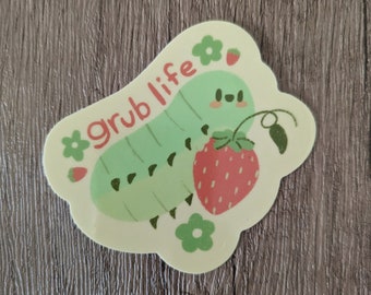 May include: A green caterpillar with a smiling face is eating a red strawberry. The text "grub life" is written in pink on a yellow background with green flowers.