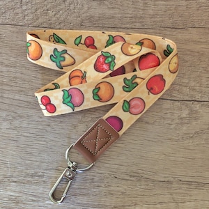 May include: A lanyard with a repeating pattern of colorful fruits, including peaches, apples, and cherries, on a light yellow background. The lanyard has a brown leather-like accent and a silver-colored metal clasp.