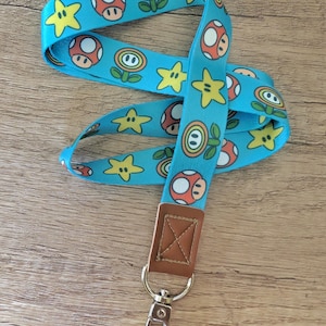 May include: A light blue lanyard featuring a repeating pattern of cartoon mushrooms, stars, and flowers. The lanyard has a brown leatherette piece with a metal clasp and ring. The lanyard is laying on a wooden surface.