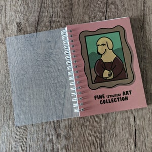 May include: A pink spiral bound notebook with a clear plastic cover. The front cover features a cartoon dog in a frame with the text "Fine (Sticker) Art Collection".