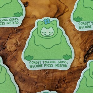 Peut inclure: Autocollants verts avec un personnage de dessin anim&eacute; dans une tenue semblable &agrave; de la mousse, une fleur bleue et le texte "FORGET TOUCHING GRASS, BECOME MOSS INSTEAD." Les autocollants sont sur une surface en bois.