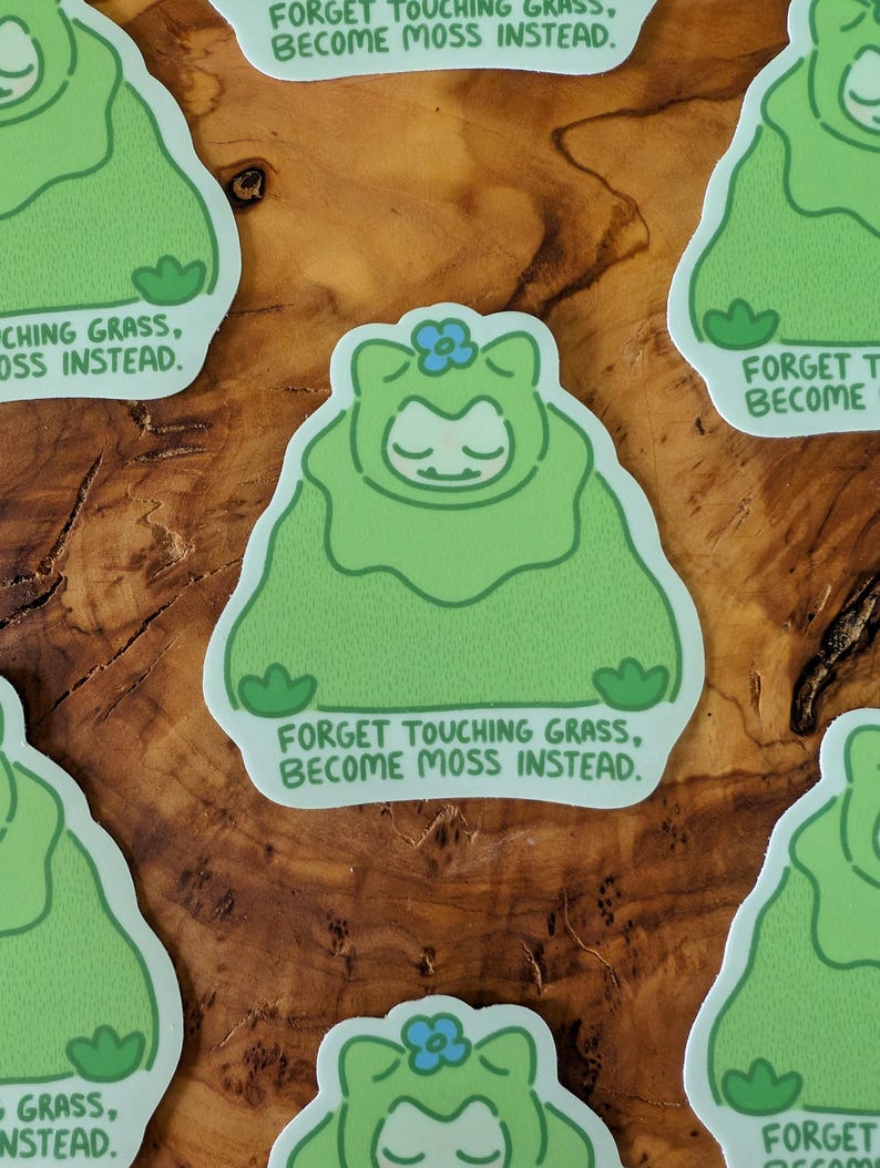 Peut inclure: Autocollants verts avec un personnage de dessin anim&eacute; dans une tenue semblable &agrave; de la mousse, une fleur bleue et le texte "FORGET TOUCHING GRASS, BECOME MOSS INSTEAD." Les autocollants sont sur une surface en bois.