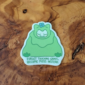 Peut inclure: Un autocollant vert repr&eacute;sentant un personnage de dessin anim&eacute; en costume de mousse avec une fleur bleue sur la t&ecirc;te. Le texte sur l'autocollant est : "FORGET TOUCHING GRASS, BECOME MOSS INSTEAD."