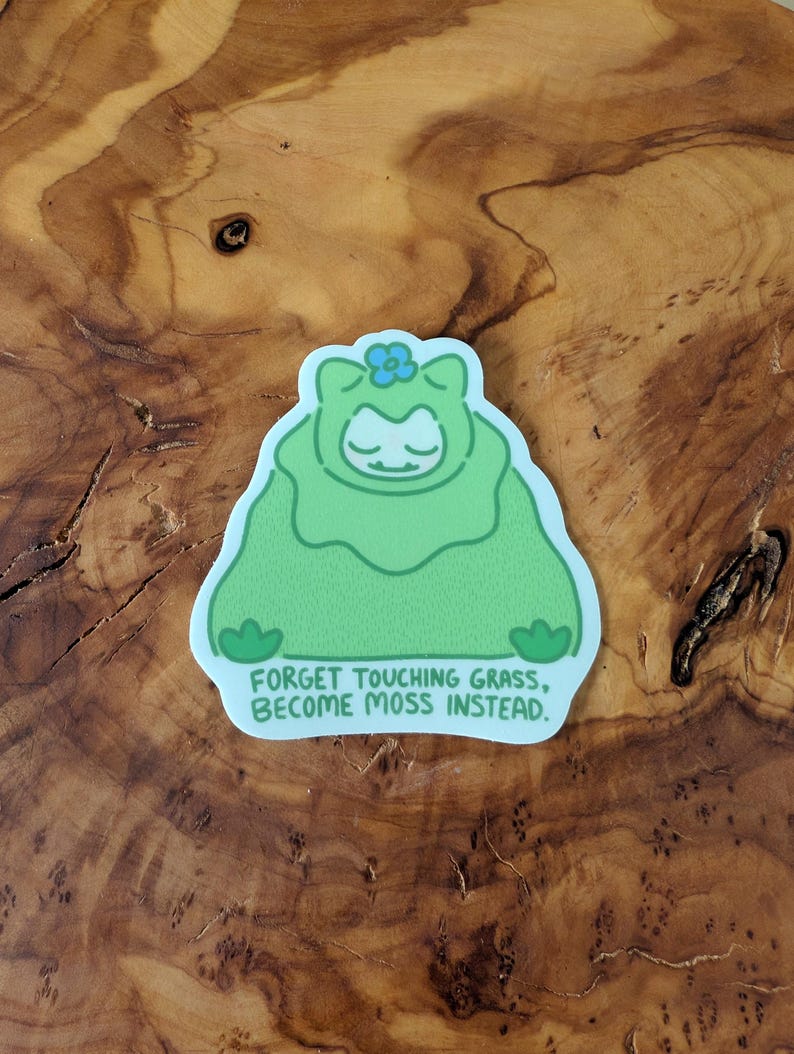 Peut inclure: Un autocollant vert repr&eacute;sentant un personnage de dessin anim&eacute; en costume de mousse avec une fleur bleue sur la t&ecirc;te. Le texte sur l'autocollant est : "FORGET TOUCHING GRASS, BECOME MOSS INSTEAD."