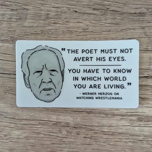 May include: A rectangular white sticker featuring a black and white illustration of Werner Herzog's face. The sticker includes a quote: "THE POET MUST NOT AVERT HIS EYES. YOU HAVE TO KNOW IN WHICH WORLD YOU ARE LIVING." - Werner Herzog on Watching Wrestlemania.