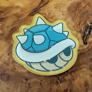 May include: A blue and white cartoon turtle shell design on a yellow background. The shell has white spikes and a white underbelly. The design is on a glossy, rounded sticker or pin.