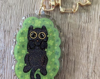 Könnte beinhalten: Ein klarer Acryl-Schlüsselanhänger mit einer schwarzen Katze mit großen, runden Augen und einem glitzernden Körper. Die Katze ist vor einem grünen Hintergrund mit Kleeblatt-Akzenten platziert. Der Schlüsselanhänger hat einen goldfarbenen Verschluss.