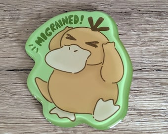 May include: A green, die-cut sticker featuring a cartoon duck with its hands on its head. The duck is light brown with a white beak and feet. The sticker has the text "MIGRAINED!" in green.