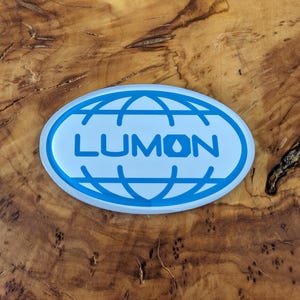 May include: An oval-shaped sticker with a light blue background and a blue outline. The word "LUMON" is printed in blue, centered within the oval, above a globe design. The sticker is placed on a wooden surface.