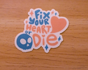 May include: A white sticker with the text "Fix your heart or Die" in pink and blue lettering. A pink heart and a blue skull are included in the design.