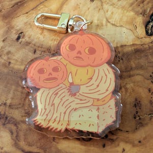 May include: A clear acrylic keychain featuring a whimsical design of two orange pumpkins. One pumpkin is holding the other. The keychain has a gold-colored clasp. The design is in shades of orange, yellow, and cream.