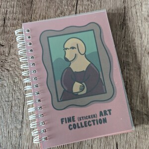 May include: A pink spiral bound notebook featuring a cartoon dog in a painting frame. The dog is wearing a purple dress and has a yellow body. The text on the cover reads "FINE (STICKER) ART COLLECTION".