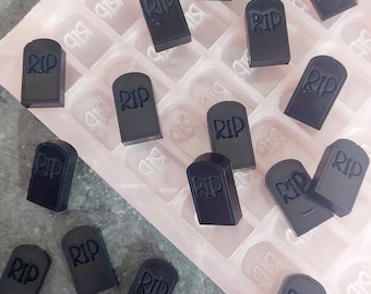 Op de afbeelding: Een close-up van een siliconen mal met meerdere holtes in de vorm van grafstenen. De mal is gevuld met zwarte hars grafstenen, elk met het woord "RIP" erop gegraveerd.
