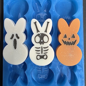 Può includere: Uno stampo in silicone blu con tre saponi a forma di coniglietto. Uno è bianco con una faccia da fantasma, un altro è bianco con un disegno di scheletro e il terzo è arancione con una faccia di zucca.