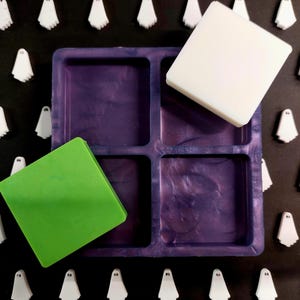 May include: A purple silicone mould with six square cavities. The mould is filled with three different coloured wax cubes: green, white and purple.
