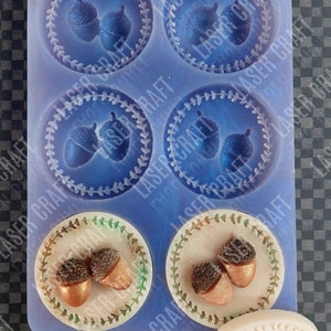May include: A silicone mould with six round cavities and one oval cavity. Each cavity features a design of two acorns with a leafy border. The mould is light blue and has the text "LASER CRAFT" printed on it.