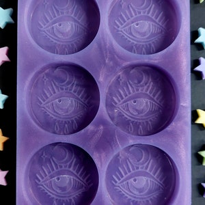 May include: Purple silicone mould with six circular cavities. Each cavity features a raised design of an eye with eyelashes, a crescent moon, and stars.