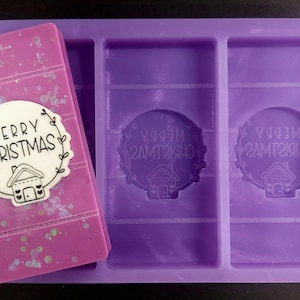 May include: Purple silicone mold with three sections. The left section shows a finished wax melt with a white circle that says "Merry Christmas" with a small house design inside. The right two sections show the mold design with the words "Merry Christmas" inside a circle with a small house design.