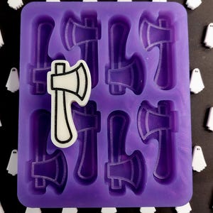 May include: A purple silicone mould with axe-shaped cavities. A white and black axe-shaped charm is placed on top of the mould. White ghost-shaped charms are scattered around the mould.