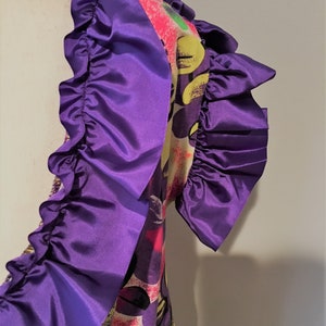 Pode incluir: Um vestido estampado colorido com fundo branco, adornado com uma guarni&ccedil;&atilde;o de babados roxos. O vestido tem uma estampa gr&aacute;fica com detalhes amarelos, rosas, verdes e pretos.