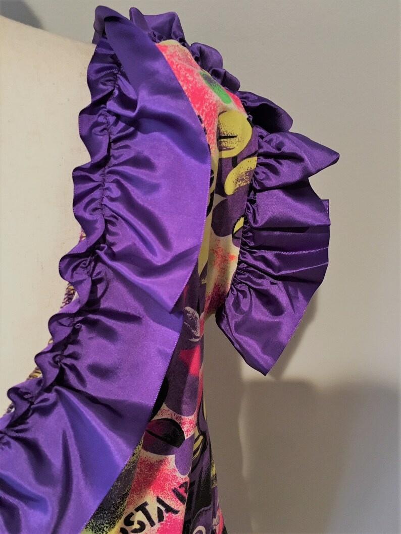 Pode incluir: Um vestido estampado colorido com fundo branco, adornado com uma guarni&ccedil;&atilde;o de babados roxos. O vestido tem uma estampa gr&aacute;fica com detalhes amarelos, rosas, verdes e pretos.