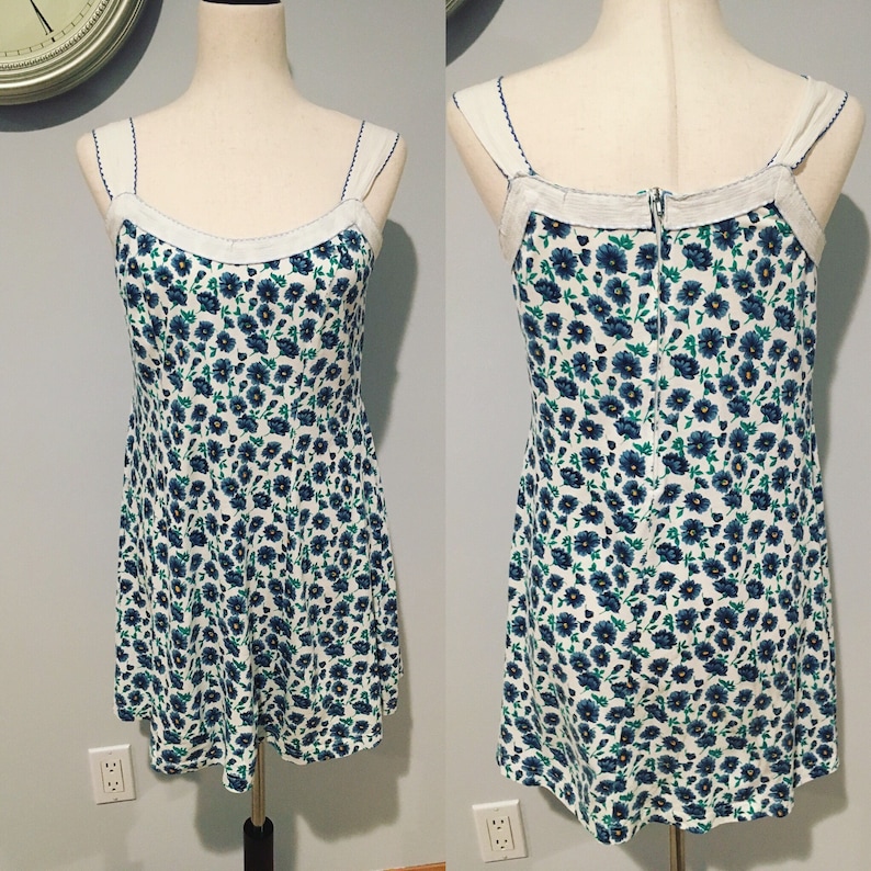 May include: A white sleeveless dress with a blue floral pattern. The dress has a scoop neckline and a fitted bodice. The skirt is A-line and falls to just above the knee.