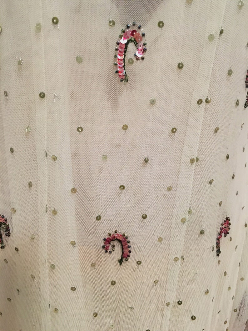May include: White sheer fabric with a repeating pattern of small green sequins and larger pink and green sequins in a curved shape.