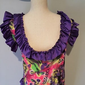 Pode incluir: Um vestido roxo e rosa com estampa floral e a palavra "COSTARICA" na frente. O vestido tem um decote com babados e mangas curtas.