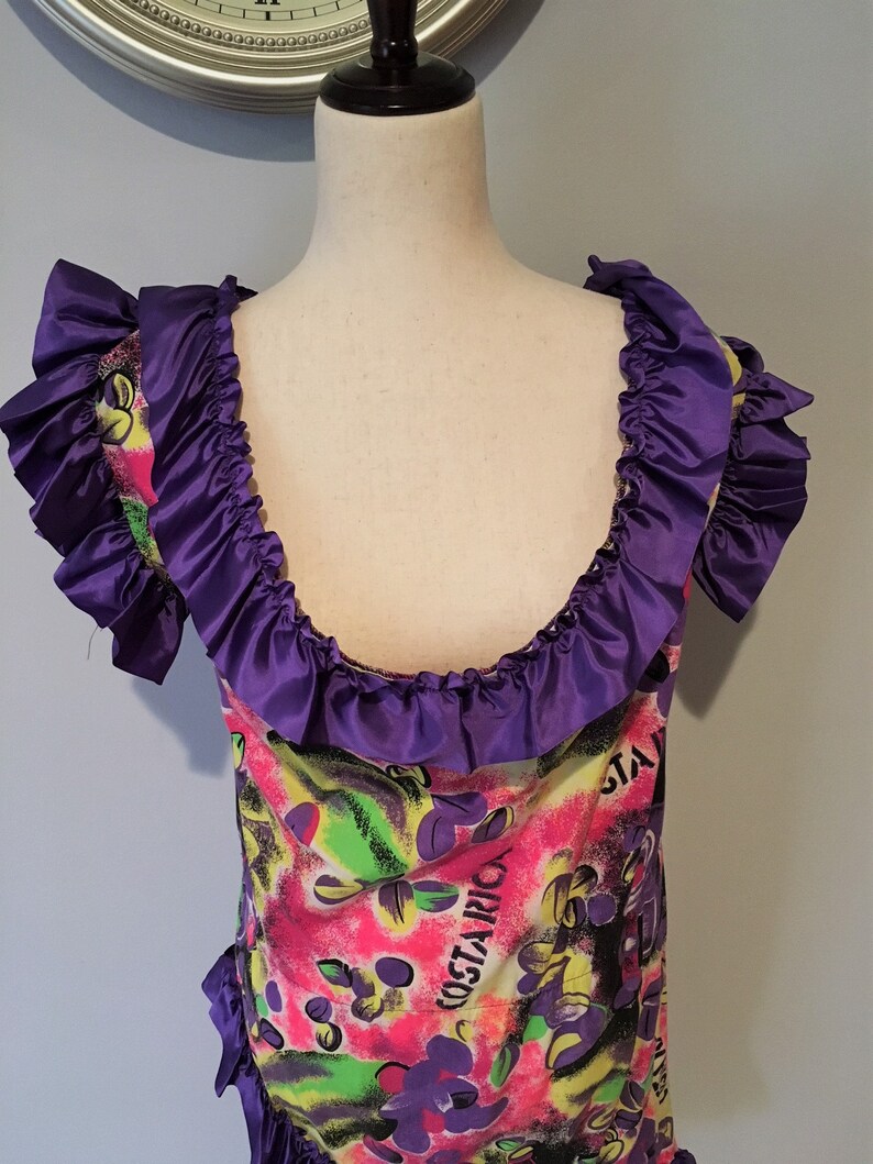 Pode incluir: Um vestido roxo e rosa com estampa floral e a palavra "COSTARICA" na frente. O vestido tem um decote com babados e mangas curtas.