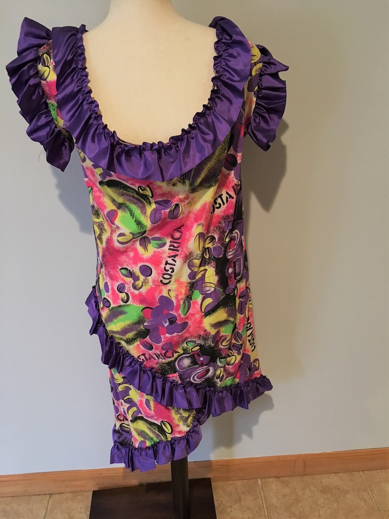 Pode incluir: Um vestido roxo e branco com decote e bainha com babados. O vestido tem uma estampa colorida com as palavras "Costa Rica" e um design floral.