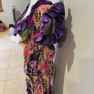 Pode incluir: Um vestido curto de manga curta colorido com uma borda de babados roxos. O vestido tem um fundo amarelo com uma estampa multicolorida de flores, folhas e as palavras "Costa Rica" e "The Night".