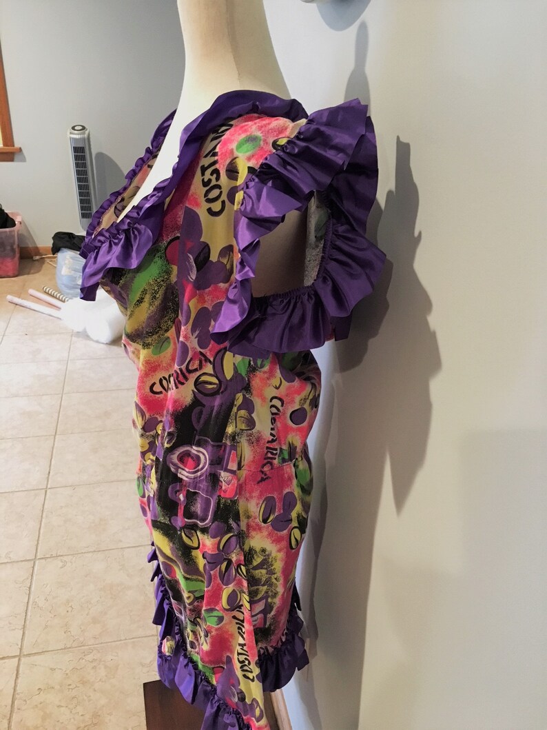 Pode incluir: Um vestido curto de manga curta colorido com uma borda de babados roxos. O vestido tem um fundo amarelo com uma estampa multicolorida de flores, folhas e as palavras "Costa Rica" e "The Night".