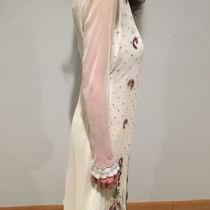 May include: A white sheer tunic with long sleeves and a beaded design. The tunic has a floral pattern with pink and green accents.