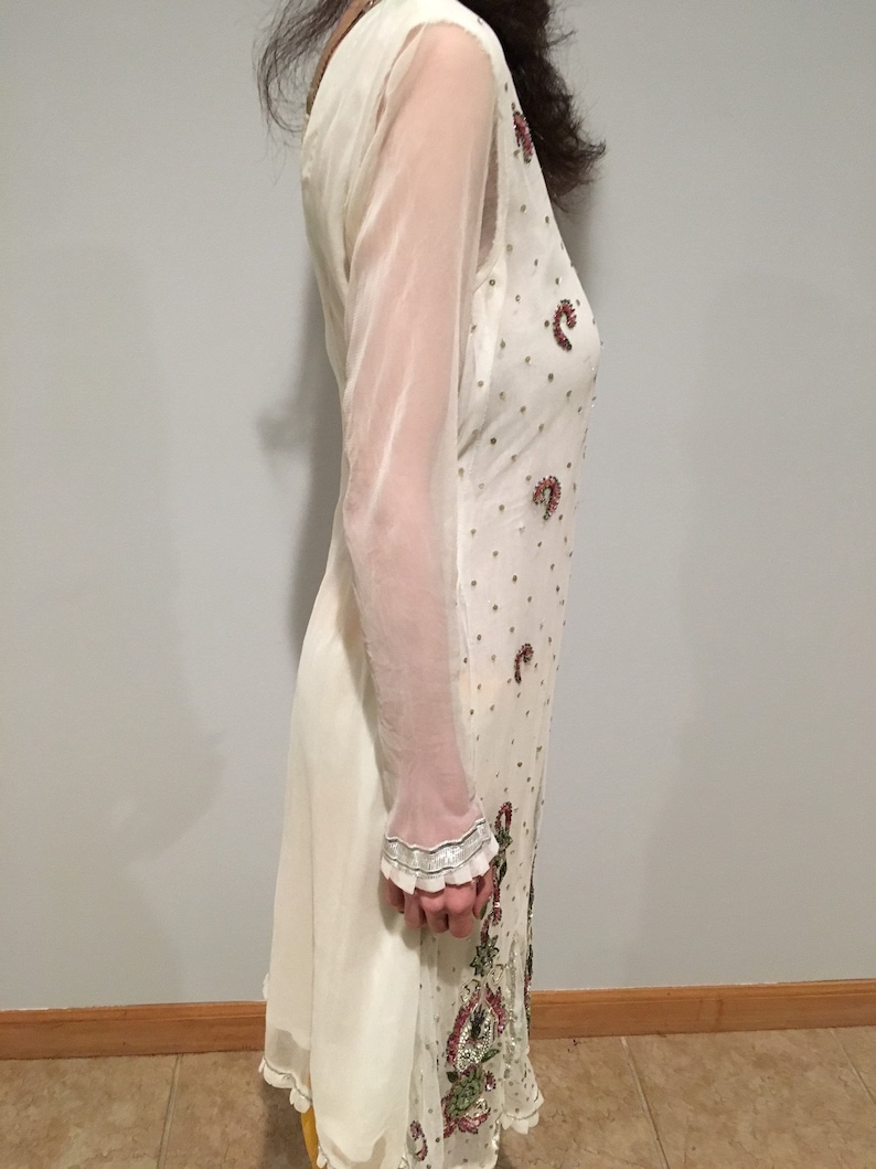 May include: A white sheer tunic with long sleeves and a beaded design. The tunic has a floral pattern with pink and green accents.