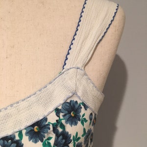 May include: A white dress with blue floral print and white straps with blue stitching. The straps are made of a textured fabric.