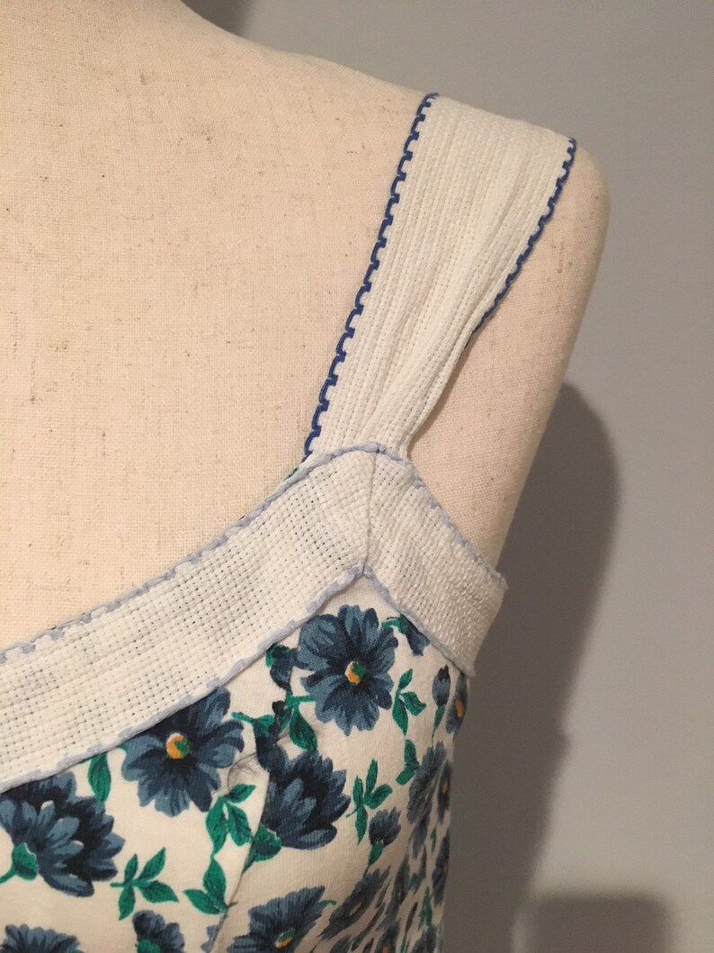 May include: A white dress with blue floral print and white straps with blue stitching. The straps are made of a textured fabric.