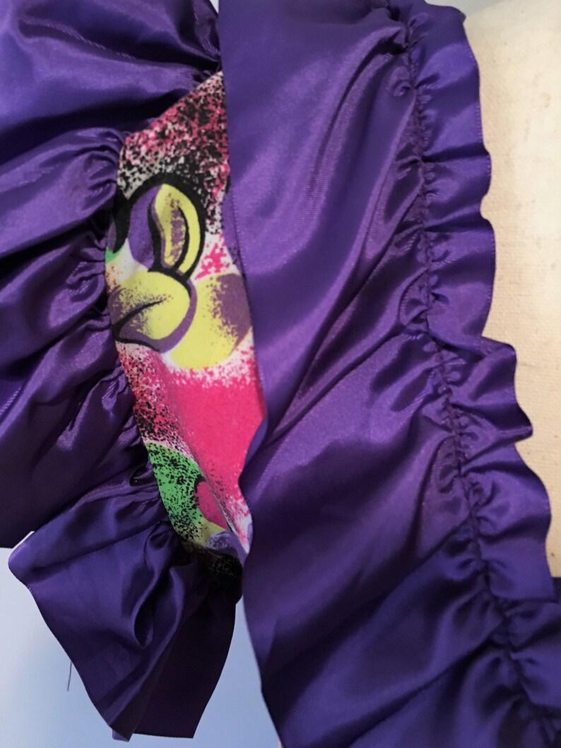 Pode incluir: Tecido de cetim roxo com uma borda franjada. O tecido tem uma estampa colorida com um personagem de desenho animado.