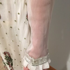 May include: White sheer long-sleeved dress with a beaded trim and a floral pattern. The dress has a ruffled cuff.