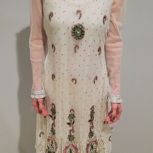 May include: A white sheer dress with long sleeves and a round neckline. The dress is decorated with pink and green floral embroidery and sequins.