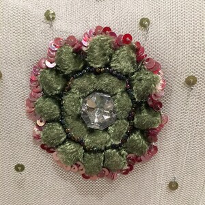 May include: A close-up of a green velvet flower with a clear rhinestone center, surrounded by pink and red sequins on a white mesh fabric.
