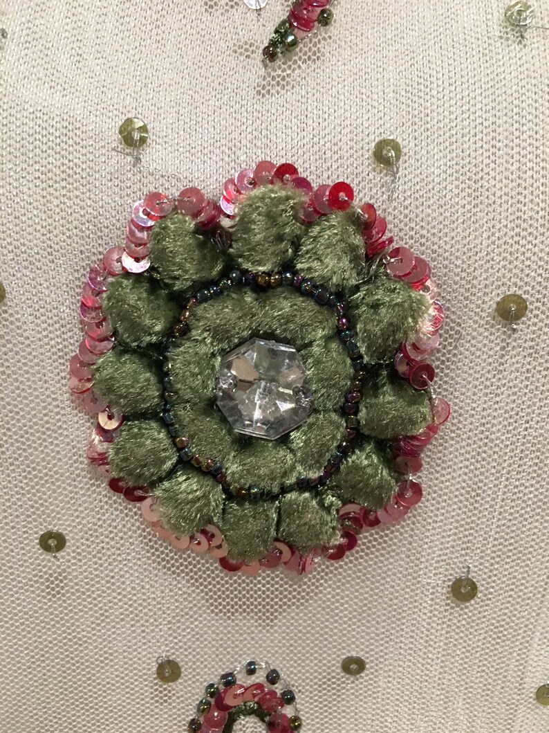May include: A close-up of a green velvet flower with a clear rhinestone center, surrounded by pink and red sequins on a white mesh fabric.