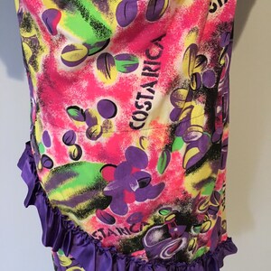 Pode incluir: Um tecido estampado abstrato colorido com uma bainha de babados roxos. O tecido apresenta um padr&atilde;o repetitivo de formas verdes, amarelas, rosas e roxas, com a palavra "COSTARICA" escrita em letras brancas.