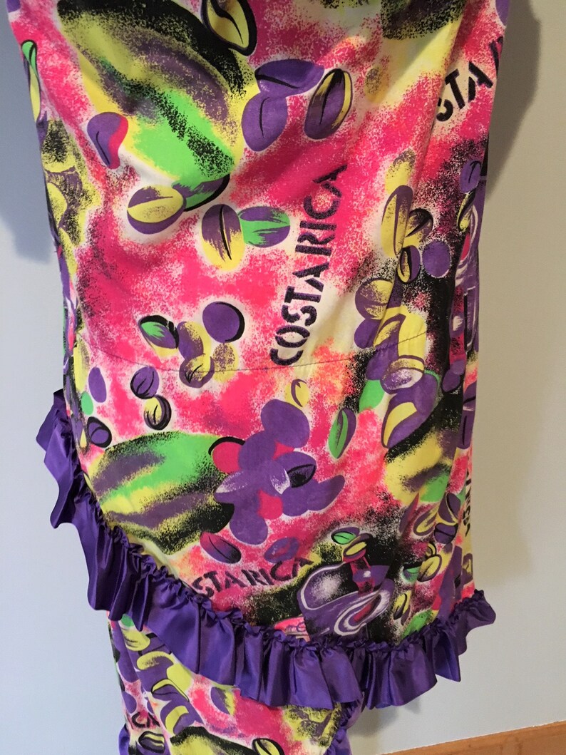 Pode incluir: Um tecido estampado abstrato colorido com uma bainha de babados roxos. O tecido apresenta um padr&atilde;o repetitivo de formas verdes, amarelas, rosas e roxas, com a palavra "COSTARICA" escrita em letras brancas.